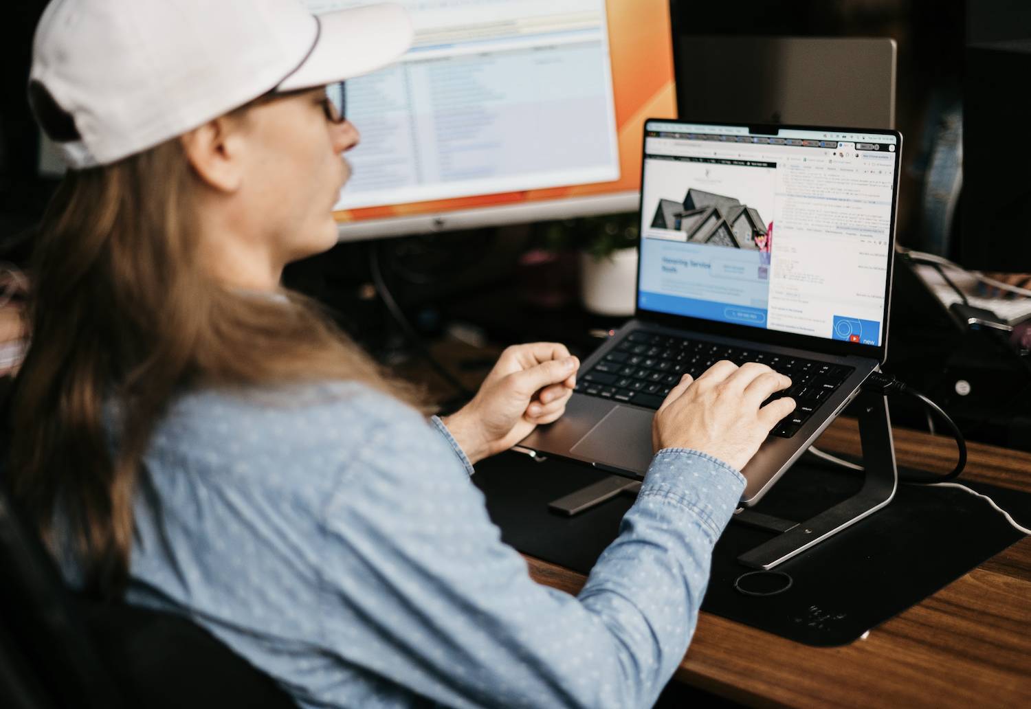 A cap-wearing individual in a blue shirt diligently types on a laptop set on a stand, focused on analyzing technical SEO data. A large monitor in the background shows a spreadsheet, indicating an office environment.