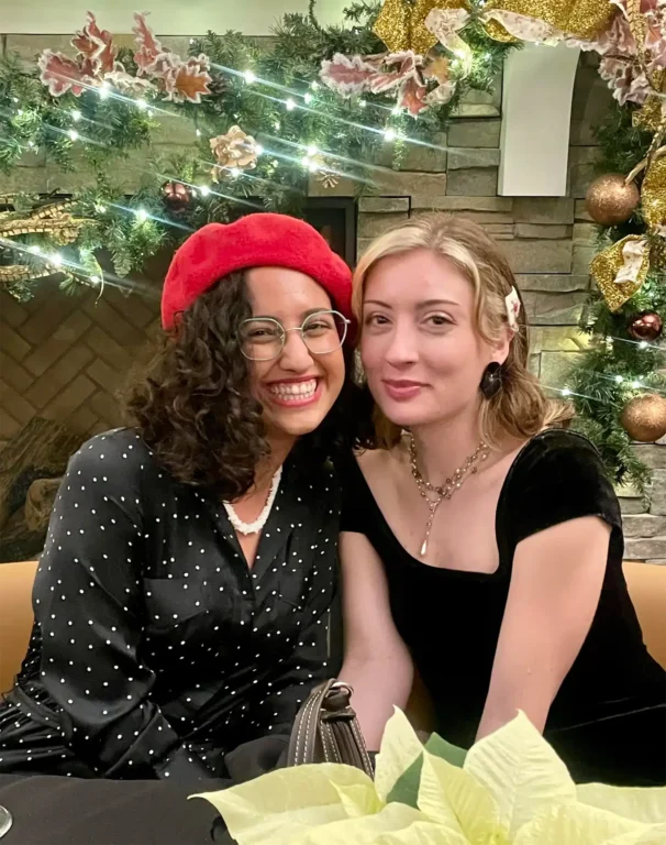 Two women, exuding joy and friendship, are seated before a festive backdrop with greenery, lights, and ornaments. One wears a red beret and polka dot blouse; the other wears a black dress. Their creative careers are highlighted by the vibrant scene, enhanced by a poinsettia in the foreground.