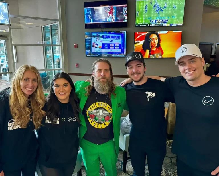 In a vibrant indoor setting, a diverse team of five professionals—three men and two women—pose confidently. One man distinguishes himself in a bright green suit while sports highlights play on the TVs behind them. Their smiles capture the essence of shared success and teamwork in their careers at Hook Agency.