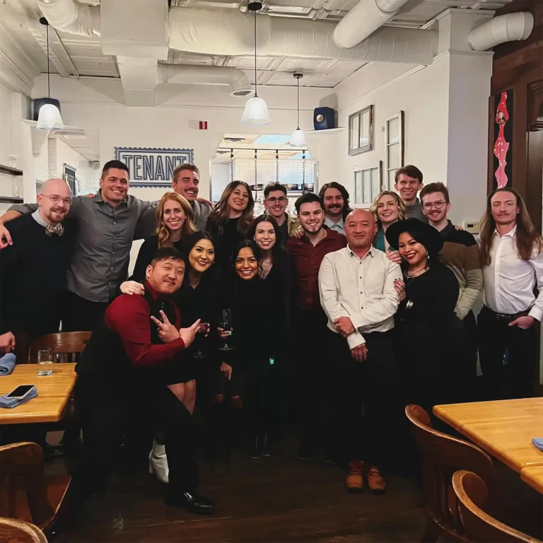 A dynamic group of 18 professionals from various industries gather inside a stylishly industrial restaurant, featuring white walls, wooden furnishings, hanging lights, and exposed ceiling pipes. They stand confidently for the camera, exuding friendly camaraderie with joyful smiles and playful gestures.
