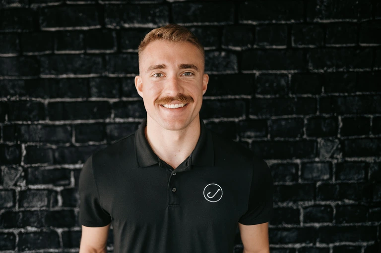 Tim Brown, with a confident smile and mustache, stands against a textured black brick wall. He is wearing a stylish black polo shirt featuring a subtle circular logo on the chest.