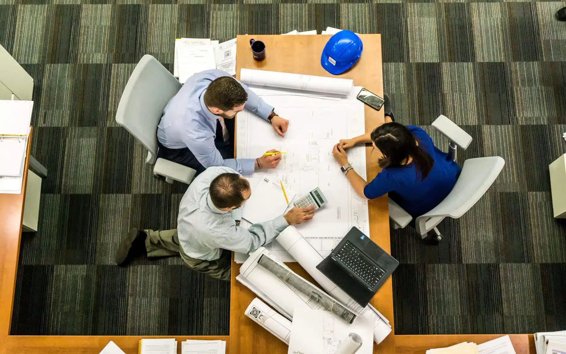 Three individuals sit at a table reviewing architectural plans, discussing potential painting strategies. A calculator, highlighter, and blue hard hat are prominently placed on the table. Nearby, a laptop and smartphone complement the scene's professional aesthetic against a striped carpet backdrop.
