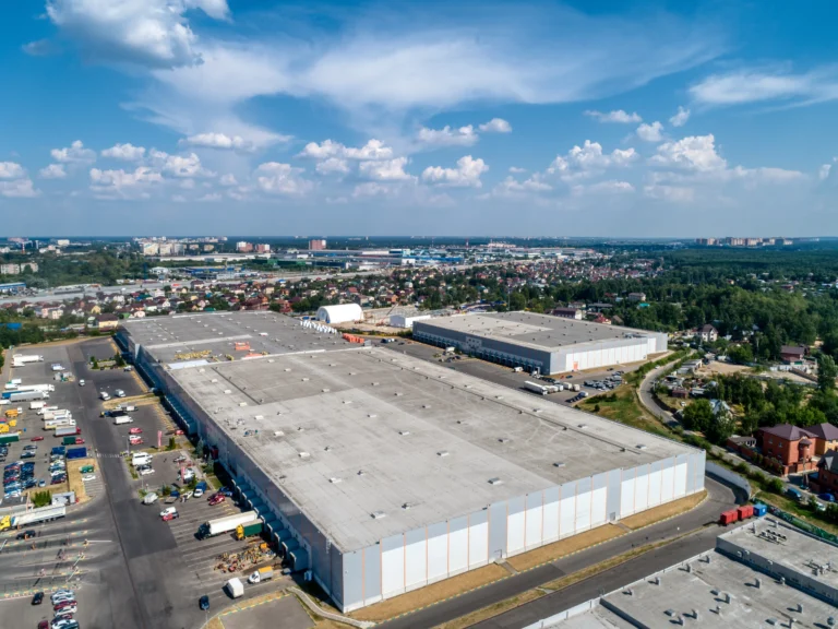 An aerial perspective showcases a large industrial warehouse featuring modern commercial roofing, encircled by a bustling parking lot with trucks and cars. This suburban setting is framed by residential areas, lush greenery, and a distant cityscape beneath a blue sky dotted with clouds.