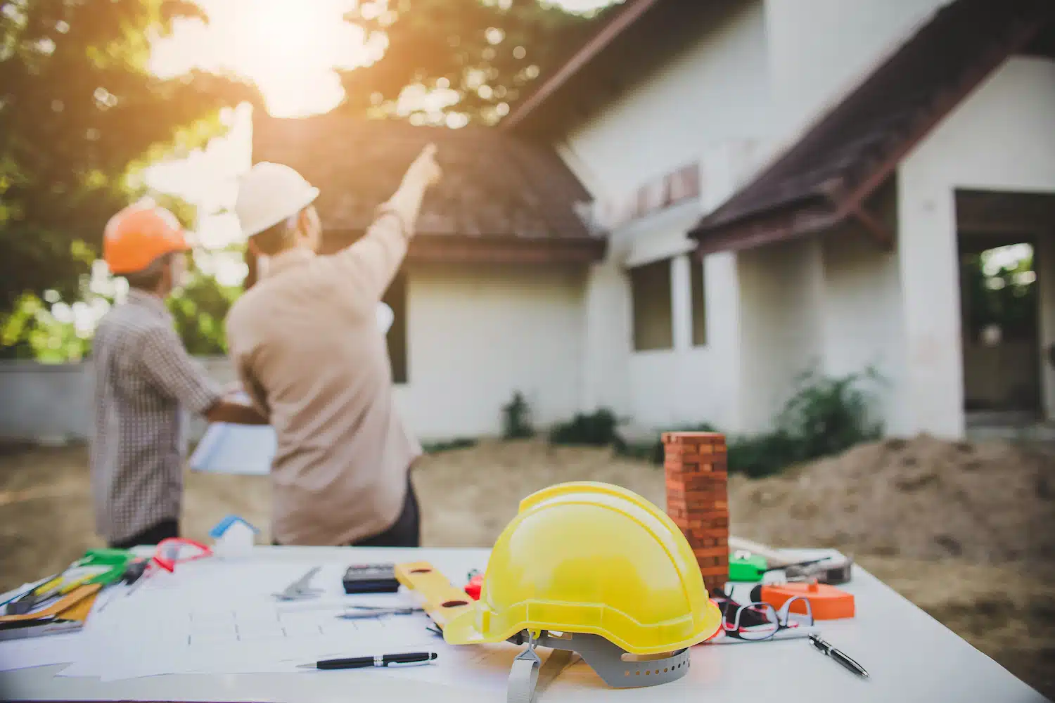 At an outdoor construction site, two workers wearing helmets discuss the progress of an unfinished house. In the foreground, a contractor's table displays plans, tools, and a yellow hard hat. Sunlight filters through the trees, illuminating their focused efforts.