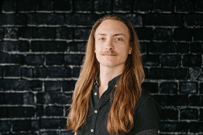 A person with long, light brown hair and a mustache, reminiscent of Tim Brown, is pictured in a black shirt adorned with white dots. Positioned against a dark brick wall, they gaze confidently at the camera, wearing a subtle smile.