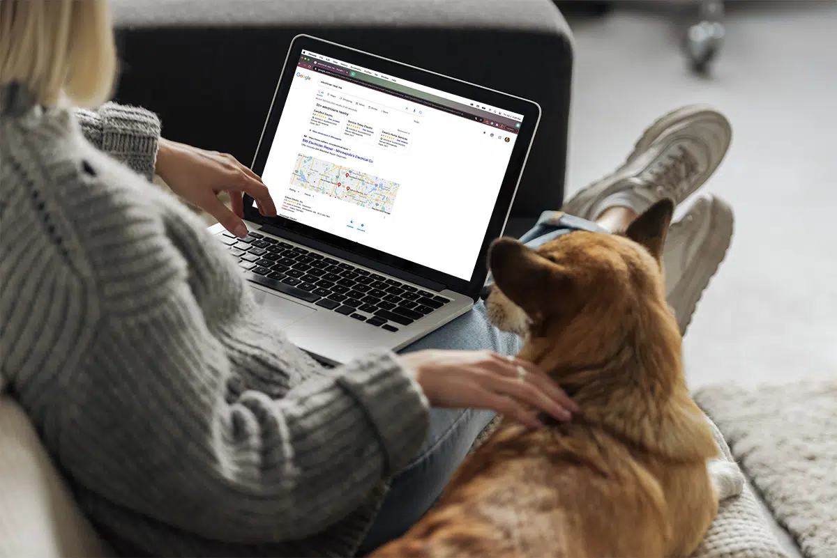 A person wearing a gray sweater sits on a couch, focused on exploring electrician marketing services on their laptop. Beside them, a brown dog watches the screen attentively. The person is relaxed, with crossed legs and white sneakers.