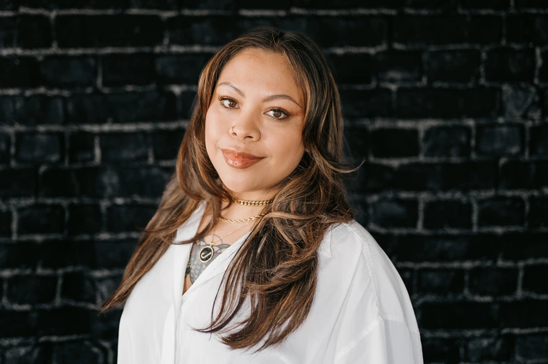 A woman with long hair, dressed in a white blouse and layered necklaces, stands against a dark brick wall. Her soft smile and serene expression echo Tim Brown's effortlessly cool style.
