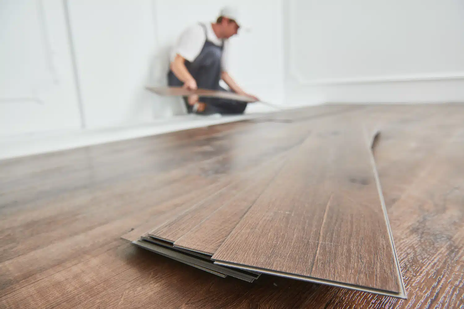 A skilled professional in work attire expertly installs wood laminate flooring against a backdrop of white walls, resembling an artful composition. Precise stacks of planks in the foreground highlight the quality craftsmanship and marketing appeal integral to exceptional service.