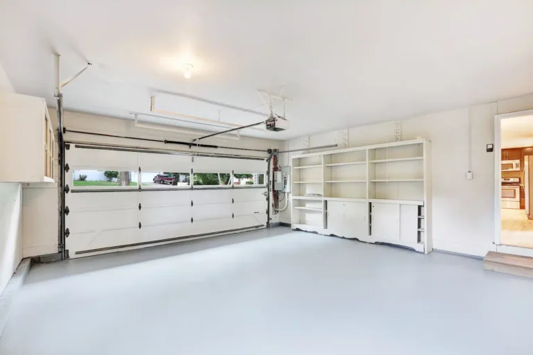 Explore our expertly transformed garage featuring a premium-coated, sleek polished concrete floor. The space is complemented by pristine white walls and ceiling, built-in side shelving units, and a closed double garage door adorned with top windows. A convenient doorway offers access to an adjoining room.