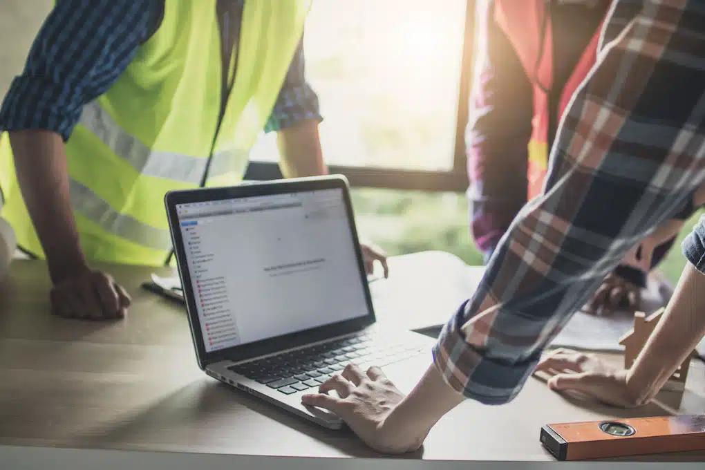 In a sunlit studio, three individuals gather around a laptop on a table. Two are clad in reflective vests, and the third wears a plaid shirt. A measuring tool positioned on the table underscores their commitment to precision work. This scene epitomizes the synergy of creativity and strategy, akin to crafting a marketing masterpiece—a hallmark of Hook Agency's approach.