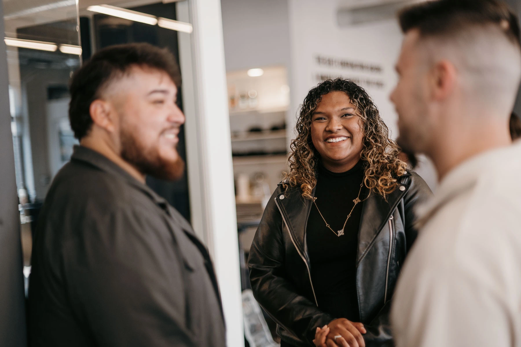 Three individuals are engaged in an indoor conversation. A person with curly hair and a black leather jacket smiles at two others: one has a beard and wears a dark jacket, while the other has short hair and sports a light shirt. The modern environment, featuring visible shelves and decor, underscores their focus on career opportunities.