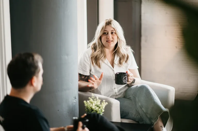A woman with long blonde hair sits in a white armchair, holding a dark mug, conversing about careers with a man in a black shirt. They are in a softly lit, modern space adorned with contemporary decor and accented by a small plant.