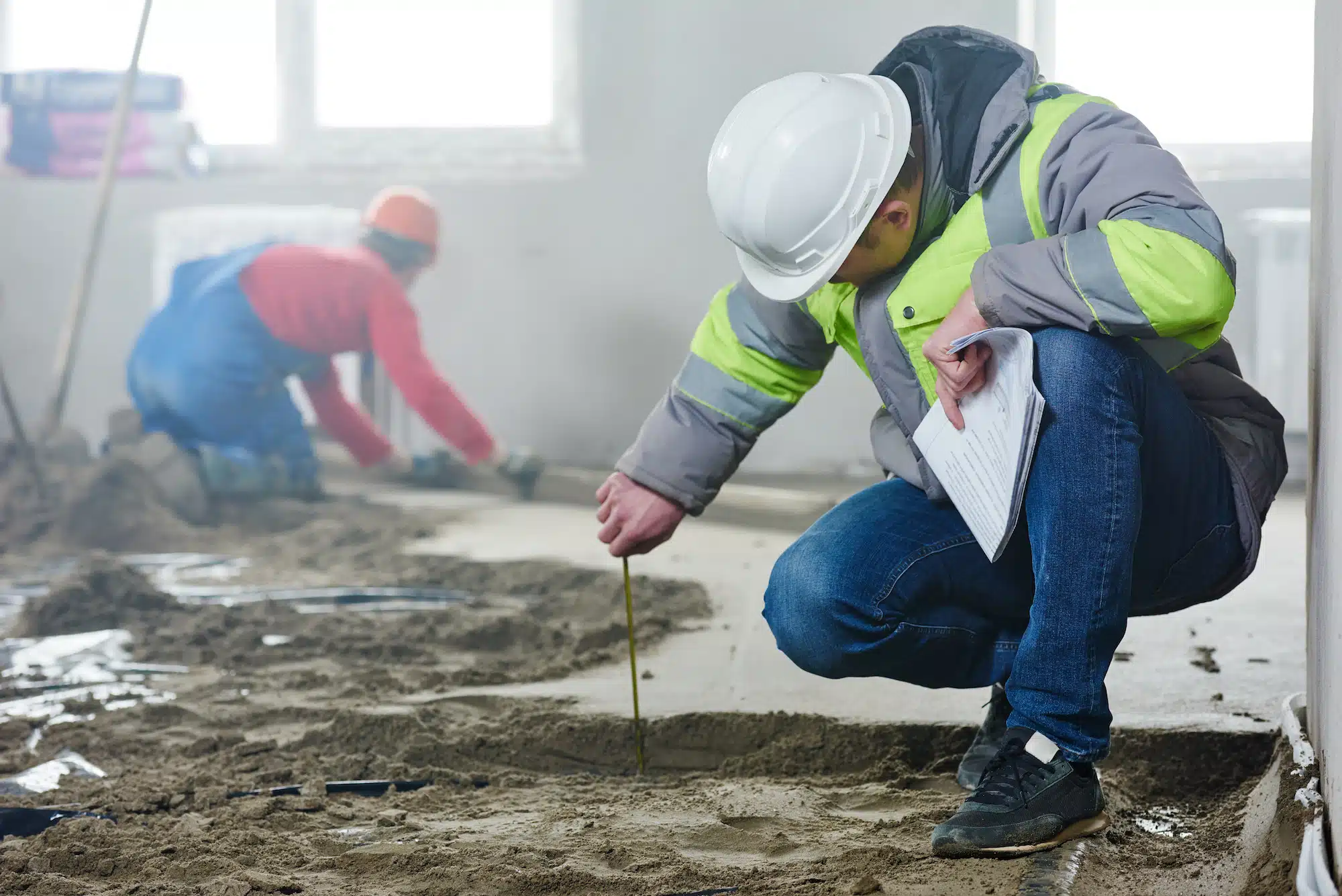 A construction worker in a hard hat and reflective jacket kneels on the sandy surface, accurately measuring with a tool while documenting on a clipboard. Nearby, another worker levels the sand, preparing for concrete pouring in the unfinished room with tall windows.
