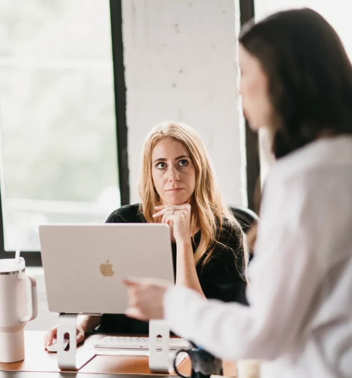 On the Hook Agency website, an image depicts two women in a bright office environment. One woman sits with her laptop, attentive and contemplative, while the other stands beside her, energetically discussing local service ads. The setting's white brick walls and large windows infuse the room with natural light.