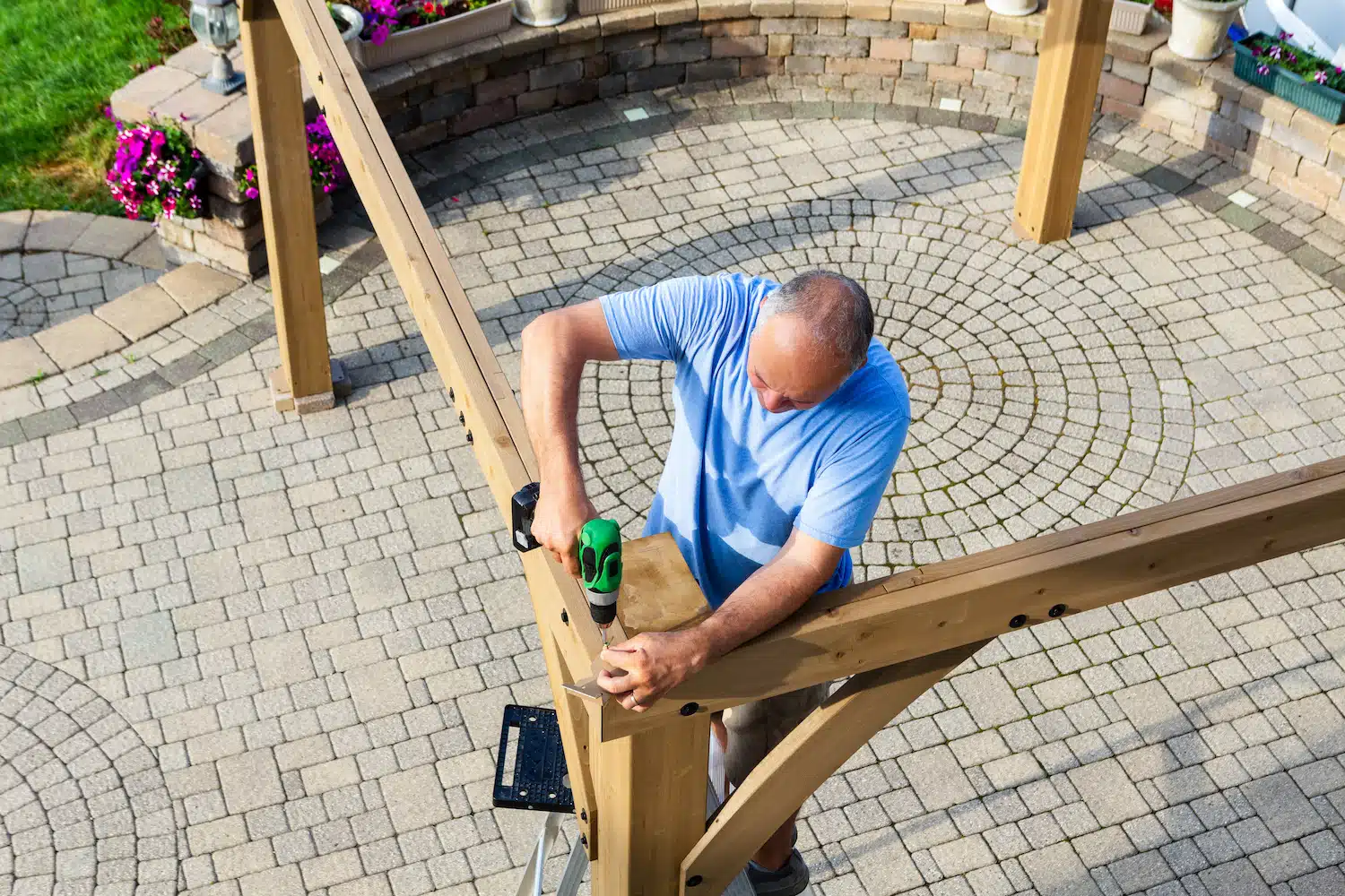 On a circular stone patio, a man in a blue shirt stands on a ladder, skillfully using a power tool to construct a wooden pergola. Sunlight filters through, casting shadows over vibrant flower beds that border the patio, enhancing the picturesque outdoor scene.