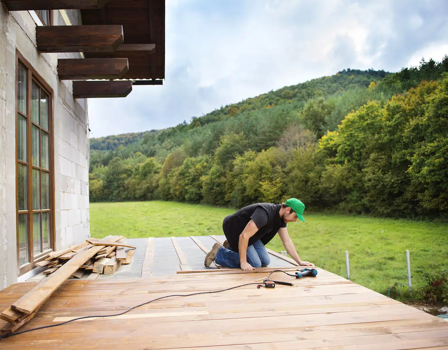 A patio contractor expertly installs wooden decking using a power tool, enveloped by lush greenery and rolling hills. Nearby, a stack of wooden planks awaits use, enhancing the tranquil yet industrious setting.