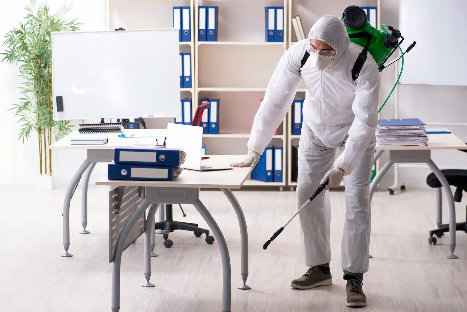On Hook Agency's website, imagine a PPC specialist as a person in full protective gear disinfecting an office space. Clad in a white hazmat suit with gloves, face mask, and goggles, they efficiently tackle digital pests using a spray wand connected to their backpack tank amidst desks and chairs.