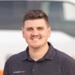 Joe Simmons, wearing a "Clean Cut" embroidered dark polo shirt, stands confidently in front of a vehicle with a blurred logo. With short hair and a friendly demeanor, he embodies professionalism and approachability.