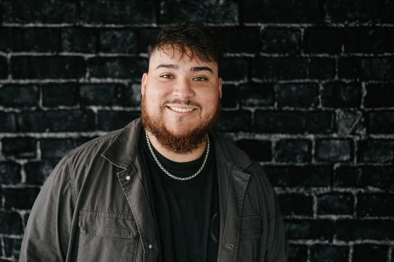 On Hook Agency's website, Tim Brown, with a beard and short hair, confidently smiles at the camera, wearing a dark shirt and jacket against a textured dark brick wall backdrop.