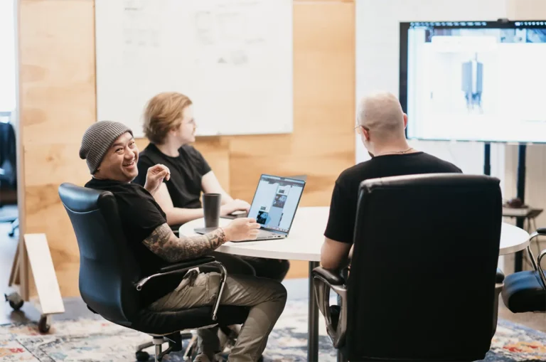 In an office setting, three professionals engage in a career-focused discussion around a table. One individual smiles with a laptop in hand, while a whiteboard and large monitor display collaborative tools in the background, promoting an inviting and team-oriented environment.