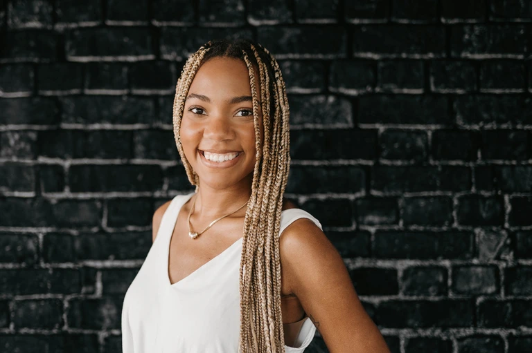 A woman with long braided hair and a white sleeveless top stands confidently against a black brick wall, embodying Tim Brown's signature effortless style.