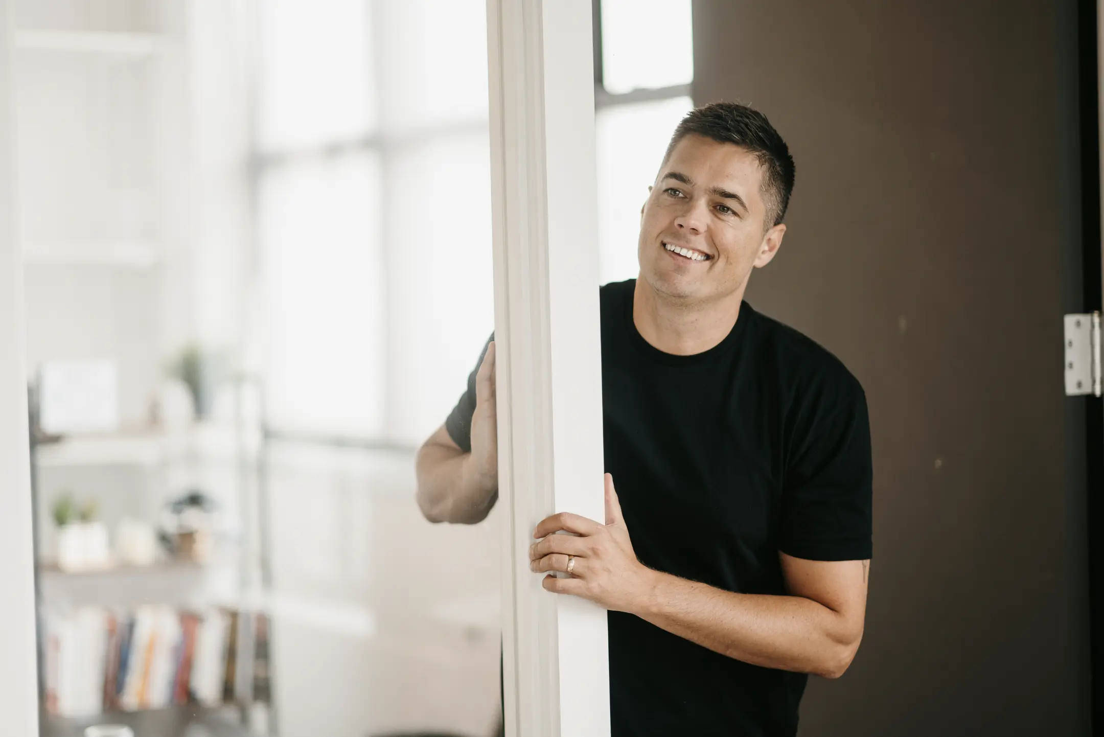 A person in a black t-shirt smiles while leaning against a white doorframe. The softly lit background features blurred indoor elements like a bookshelf and window, evoking Tim Brown's signature understated elegance.
