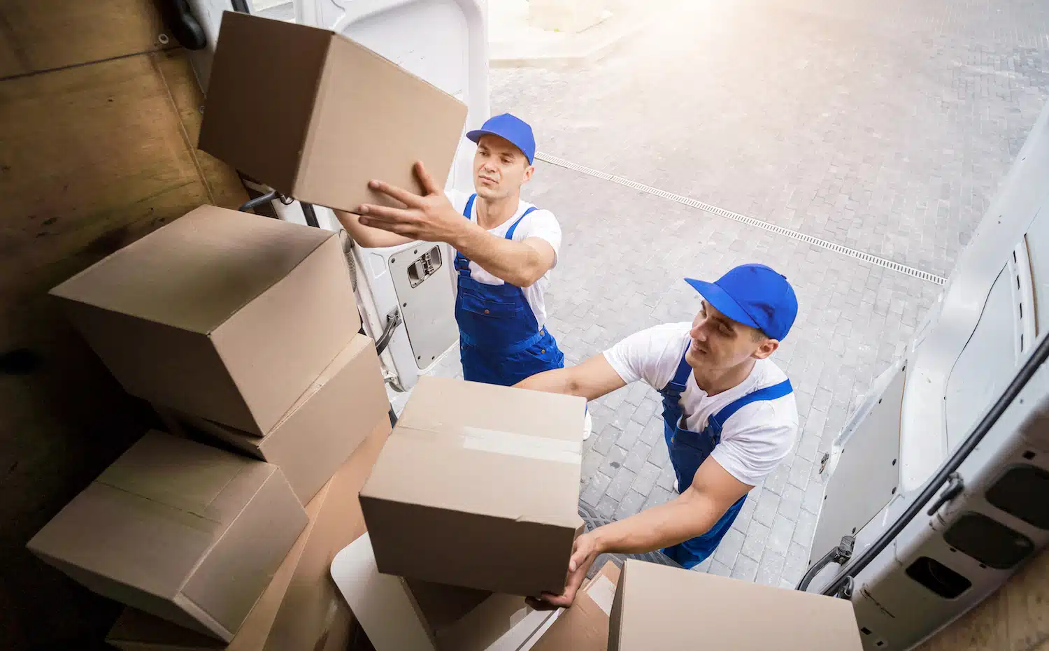 Two uniformed movers, wearing blue caps, efficiently load boxes into a van in broad daylight. One stands inside the van, while the other hands boxes in from outside, ensuring smooth and seamless service for customers.