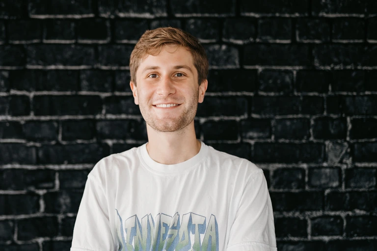 Tim Brown stands confidently against a black brick wall, sporting short hair and a light beard. He smiles at the camera in a white graphic t-shirt.