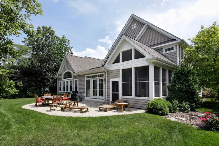A modern suburban house with large windows, a screened porch, and a gabled roof. The backyard features a stone patio, crafted by expert patio contractors, with wooden chairs and tables, surrounded by a well-maintained lawn and trees under a partly cloudy sky.