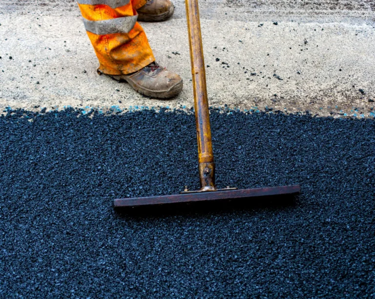 On Hook Agency's website, an image captures a paving worker in orange reflective pants and brown boots expertly smoothing fresh black asphalt with a wooden-handled tool. Above, a completed section of road showcases its flawless texture, effectively illustrating the superior quality of seamless roadwork.