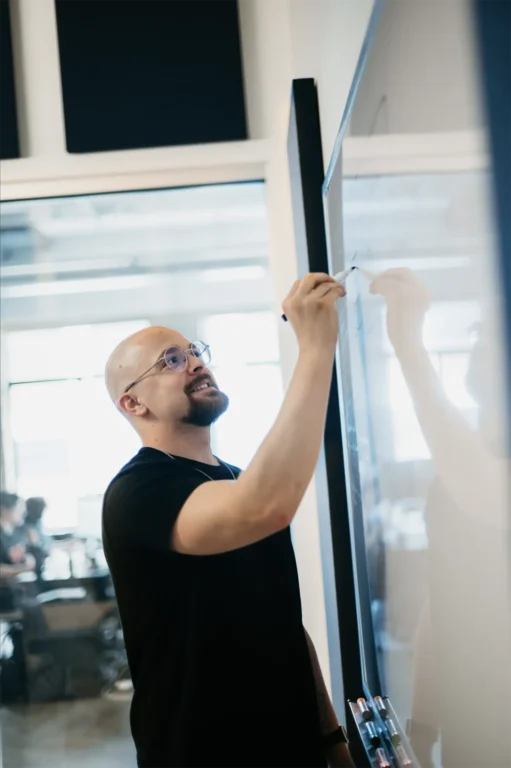 A bearded man in a black shirt writes intently on a glass board in an office, exuding focused creativity. In the blurred background, team members work diligently at their desks, enhancing the atmosphere of innovation and productivity.