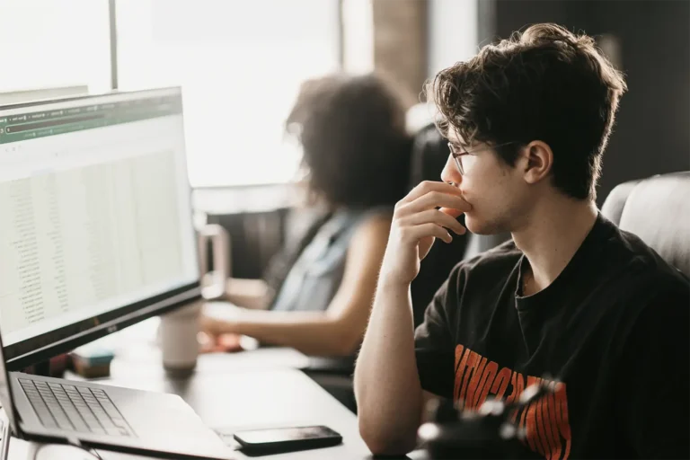 A young professional, wearing glasses, meticulously examines a spreadsheet on his computer screen. Behind him, a colleague is engaged in her tasks at another desk. This scene embodies the concentrated and dynamic atmosphere of our office at Hook Agency.