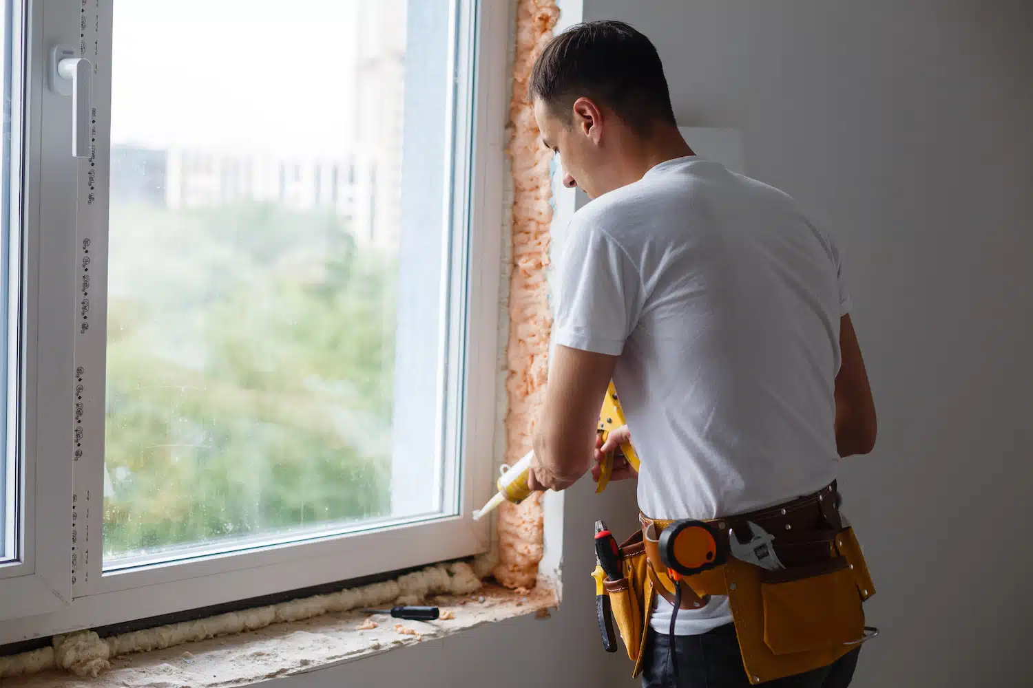 A skilled professional in a white shirt and tool belt expertly seals a window edge with a caulking gun during a remodeling project. This sealed window not only frames an elegant view of trees and buildings but also enhances home efficiency and aesthetics, aligning with SEO-friendly upgrade practices. Remodeling SEO Services and Remodeling Company Names