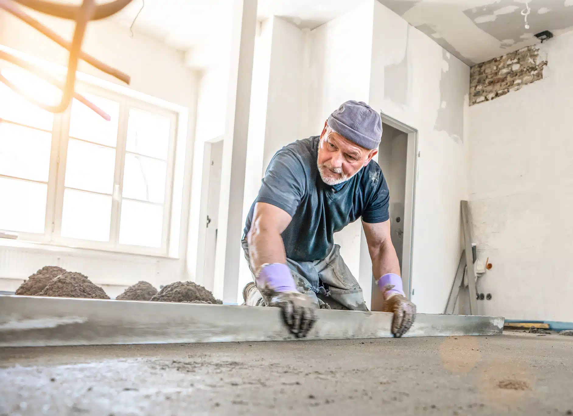 A skilled professional kneels on the floor, precisely leveling concrete in a sunlit room, underlining the transformation at hand. Piles of concrete indicate ongoing remodeling efforts, illuminated by streaming sunlight through the windows.