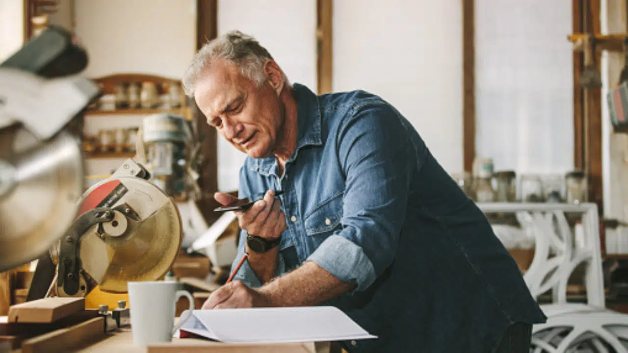 HVAC link building, Roofing Link Building - An experienced craftsman in a denim shirt leans over his workbench, taking notes on roofing techniques as he discusses link-building strategies over the phone. Nearby, a circular saw and cup are set against the rich backdrop of tools and materials in his workshop.