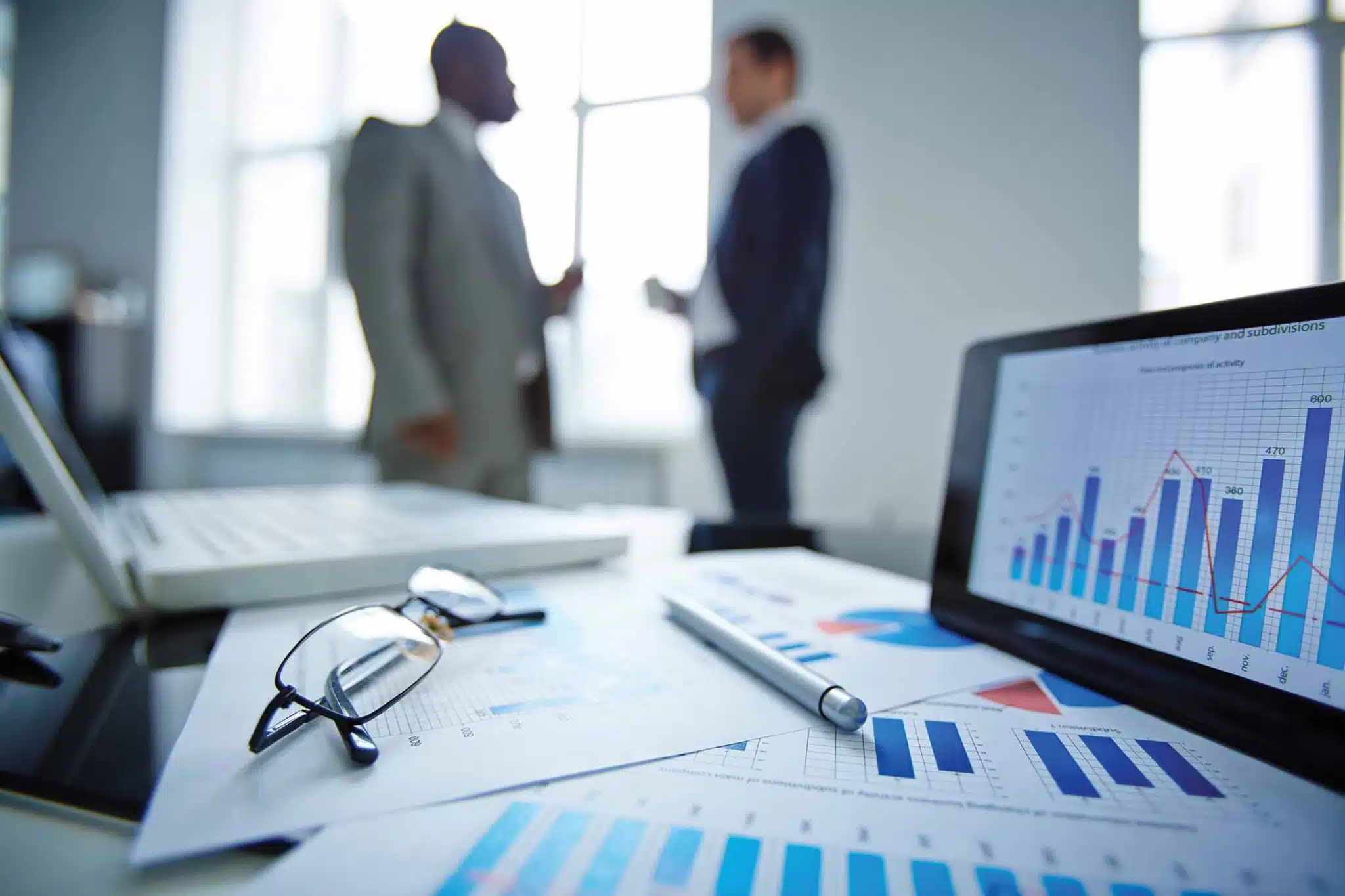 Roofing keywords, roofing private equity On Hook Agency's website, a desktop scene features glasses, papers, and a pen alongside vibrant roofing SEO charts and graphs. In the background, two professionals in business attire converse in a brightly lit office.