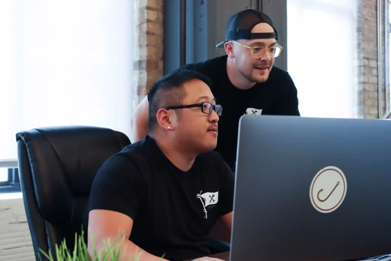 In an industrious office with large windows and brick walls, two team members in black shirts collaborate on commercial roofing SEO strategies at a desk with a large monitor. One is seated while the other stands, both intently focused on their work.