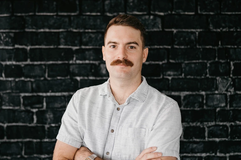 Tim Brown, sporting a mustache and wearing a light-colored short-sleeve button-up shirt, stands confidently with arms crossed against a dark brick wall.