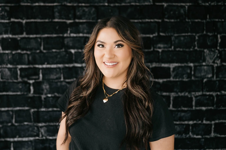 A woman with long, wavy brown hair stands confidently in front of a black brick wall, embodying Tim Brown's iconic urban style. She wears a black top and accessorizes with a heart pendant gold necklace.