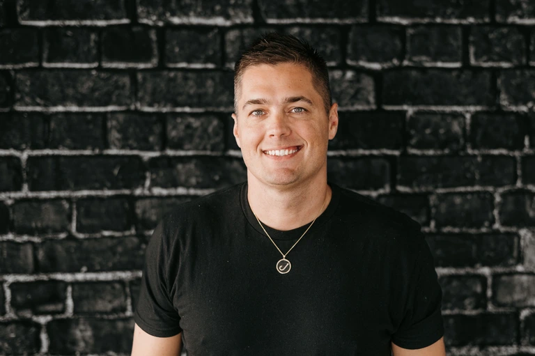 Tim Brown, in a black shirt and circular pendant necklace, smiles confidently against a black brick wall background on Hook Agency's website.