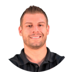 A professional man with short brown hair, a trimmed beard, and a black collared shirt smiles confidently against a clean white background.