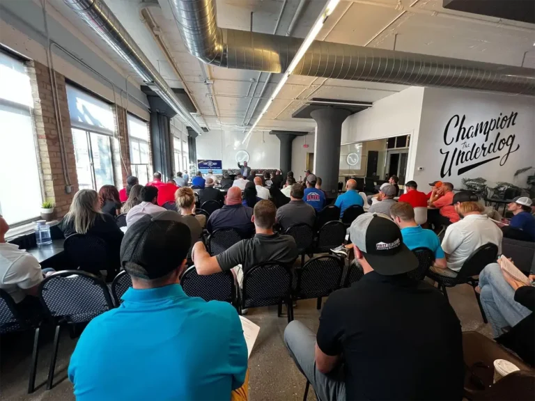 In a spacious, industrially decorated room with expansive windows, attendees focus intently on a speaker discussing career paths. The prominent "Champion Underdog" slogan on the wall serves as a powerful reminder: pursue your goals relentlessly.
