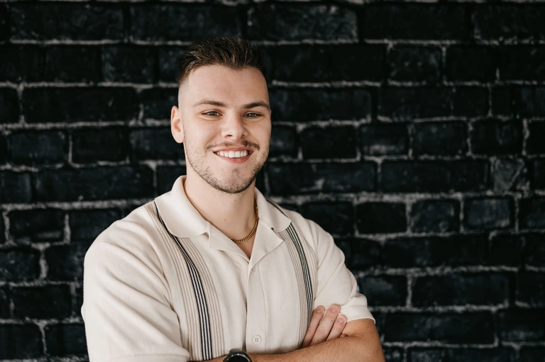 Vincenzo Colosimo, confidently posed with arms crossed, stands against a dark brick wall. He wears a light-colored shirt paired with suspenders and a watch.